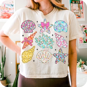 May include: A white t-shirt with a colorful seashell and starfish design. The design features a variety of seashells, starfish, and a pink bow.