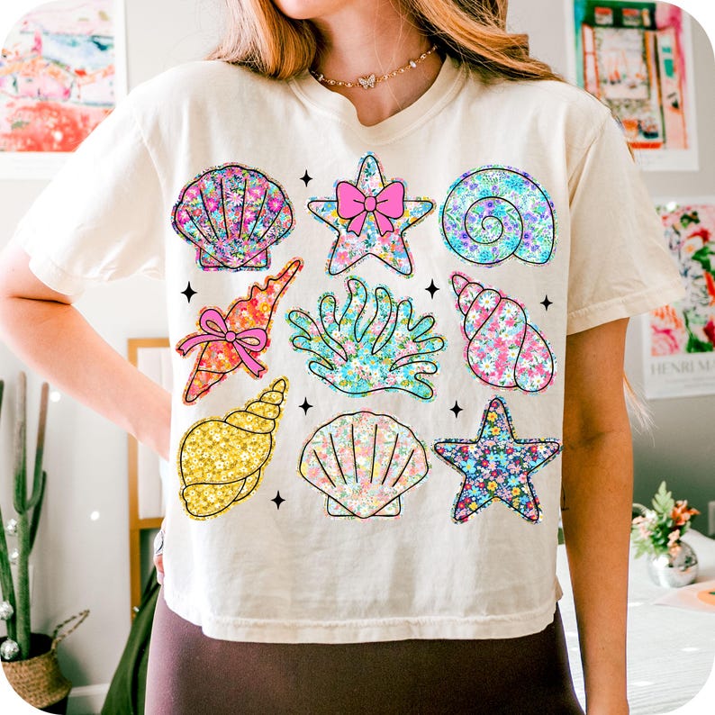 May include: A white t-shirt with a colorful seashell and starfish design. The design features a variety of seashells, starfish, and a pink bow.