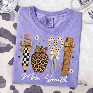 May include: A light purple t-shirt featuring an embroidered design of a checkered pencil, a leopard print apple, scissors with a polka dot pattern, and a wooden ruler. Each item is accented with a tan bow. The text Mrs. Smith is embroidered below.