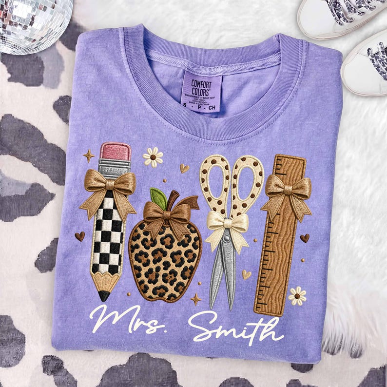 May include: A light purple t-shirt featuring an embroidered design of a checkered pencil, a leopard print apple, scissors with a polka dot pattern, and a wooden ruler. Each item is accented with a tan bow. The text Mrs. Smith is embroidered below.