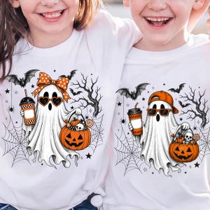 May include: Two white t-shirts featuring a Halloween graphic. One ghost wears a polka-dot bow and heart-shaped sunglasses, holding a pumpkin bucket and a drink. The other ghost wears a baseball cap and sunglasses, also holding a pumpkin bucket and drink.