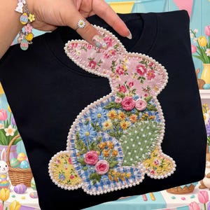 May include: Black sweatshirt featuring a patchwork bunny design. The bunny is adorned with floral patterns, embroidered details, and a pearl bead outline. The design incorporates pink, blue, yellow, and green fabrics, creating a festive Easter theme.