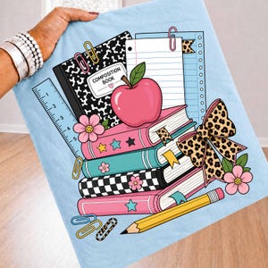 May include: A light blue t-shirt featuring a graphic illustration of school supplies including a black and white composition book, a stack of colorful books, a red apple, a yellow pencil, a ruler, paper clips, and a leopard print bow.