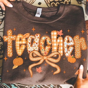 May include: A brown sweatshirt with the word "teacher" in a decorative font. The letters are filled with fall-themed patterns, including pumpkins, leaves, and floral designs. A large bow and star accents complete the design.
