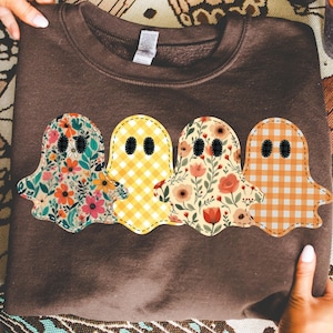 Op de afbeelding: Een bruine sweatshirt met vier spookapplicaties. Elke geest heeft een uniek bloemen- of ruitpatroon in roze, oranje, geel en groen. De geesten hebben zwarte knoopogen en zijn op de sweatshirt genaaid.