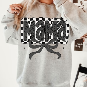 May include: A light gray sweatshirt featuring the word "Mama" in a black leopard print design, set against a black and white checkered background. A black bow is below the text, with small star accents.