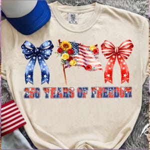 May include: A cream-coloured t-shirt with patriotic designs. It features a blue bow with white stars, a floral American flag, and a red bow with white stars. The text "250 YEARS OF FREEDOM" is displayed below the graphics.