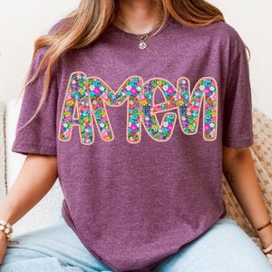 May include: A purple t-shirt with the word "Amen" in large, white letters outlined in pink. The letters are filled with a colorful floral pattern.