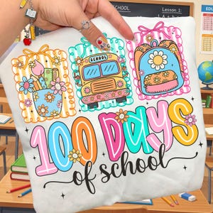 以下が含まれることがあります： 「100 Days of School」を祝うカラフルなグラフィックがプリントされた白いTシャツ。デザインには、スクールバス、バックパック、花瓶の花が含まれており、すべて装飾的なボーダーで囲まれています。テキストは遊び心のあるマルチカラーフォントです。