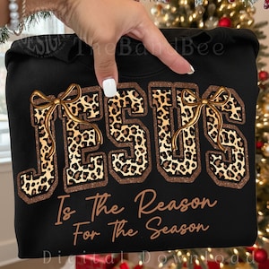 May include: Black sweatshirt featuring the word "JESUS" in leopard print letters with gold ribbon accents. The phrase "Is The Reason For The Season" is printed below in a brown script font. The sweatshirt is held up by a hand.