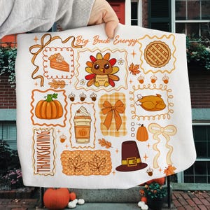 Könnte beinhalten: Ein weißes, dekoratives Tuch zum Thema Thanksgiving mit verschiedenen orangefarbenen und braunen Illustrationen. Designs umfassen einen Kürbis, Truthahn, Kuchen, Kaffee und die Texte "Big Buck Energy" und "Thanksgiving".