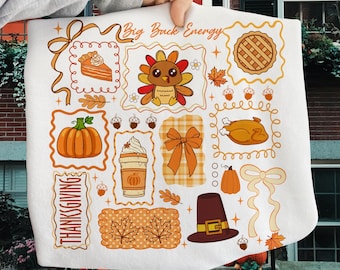 Pode incluir: Um tecido decorativo branco com ilustrações temáticas de Ação de Graças em laranja e castanho. Os desenhos incluem uma abóbora, peru, torta, café e os textos "Big Buck Energy" e "Thanksgiving".