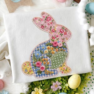 May include: A white fabric square features a patchwork bunny design. The bunny is adorned with floral patterns in pink, blue, yellow, and green, outlined with a pearl-like trim. The design includes embroidered flowers and a polka-dot accent, perfect for Easter.