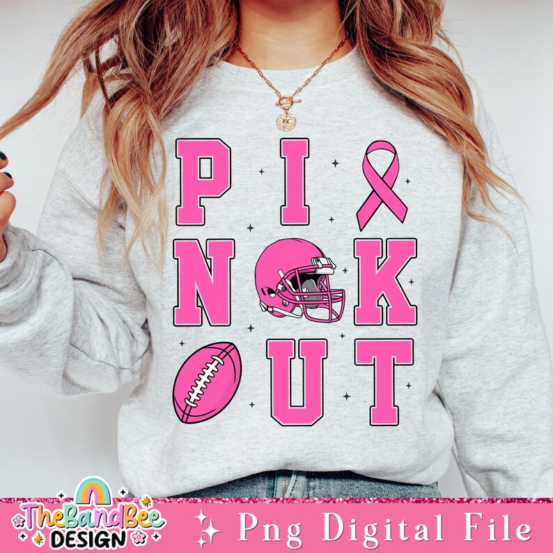 K&ouml;nnte beinhalten: Rosa-wei&szlig;es Sweatshirt mit dem Text "PINK OUT" in rosa Buchstaben. Der Text ist in einem Block mit einem Fu&szlig;ball, einem Footballhelm und einem rosa Band angeordnet.