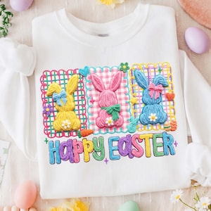 May include: White sweatshirt with an embroidered design of three Easter bunnies in yellow, pink, and blue, each with a bow and flower. The words "HAPPY EASTER" are written in colourful letters below the bunnies. The design is framed with a colourful grid.
