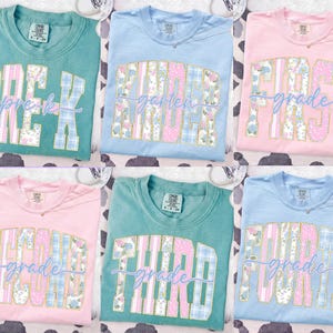 Può includere: Sei magliette dai colori pastello con testo di livello scolastico. Le magliette sono azzurre, rosa e verde acqua. Ogni maglietta ha un livello scolastico, come "Pre-K" o "First Grade", in un design floreale e a quadri.