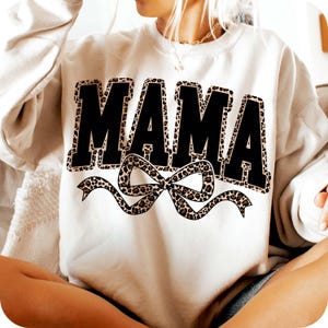 May include: Cream-colored sweatshirt with the word "MAMA" in large, black, block letters. The letters are outlined with a leopard print pattern. A leopard print bow is centered below the word.