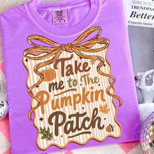May include: A lilac Comfort Colors t-shirt featuring a decorative design with the text "Take me to The Pumpkin Patch." The design includes a pumpkin, leaves, and a ribbon. The shirt is folded.