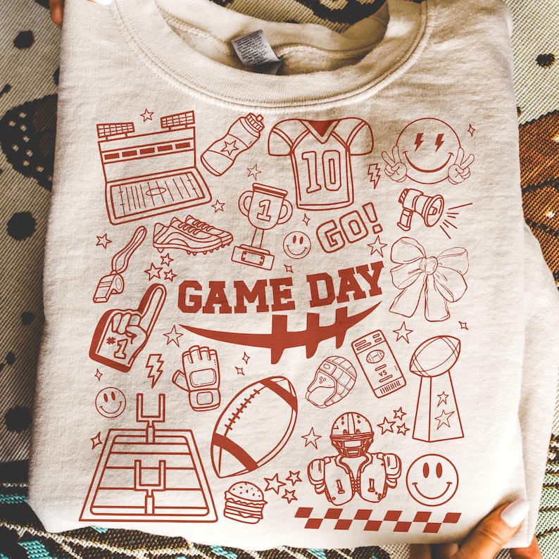 May include: A light beige sweatshirt with a red graphic design featuring football-related icons. The design includes a football field, helmet, football, and the words "GAME DAY".