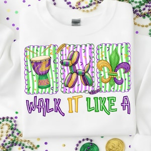 Puede incluir: Sudadera blanca con un diseño gráfico colorido que presenta un tambor, un animal de globo y una flor de lis. El texto "WALK IT LIKE A" está impreso en púrpura, verde y amarillo. El diseño tiene un tema de Mardi Gras.
