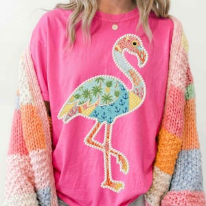 Może przedstawiać: Różowa koszulka z kolorowym wzorem flaminga z różnymi wzorami, w tym palmami i kotwicami. Flaming jest obrysowany białymi koralikami. Wielokolorowy sweter jest noszony na koszulce.