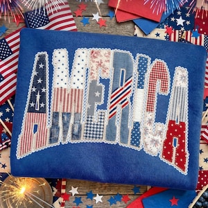 May include: A blue sweatshirt featuring the word "AMERICA" in a patchwork design. The letters are crafted from fabrics with star, stripe, and floral patterns. The image is set against a backdrop of American flags and festive decorations.