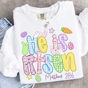 May include: White sweatshirt with the words "He is Risen" in colorful, patterned letters. The design includes Easter-themed graphics like bunnies, eggs, and a carrot. The text "Matthew 28:6" is printed below the main text.
