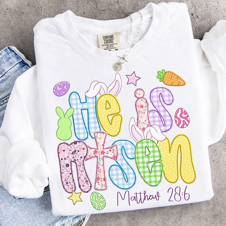May include: White sweatshirt with the words "He is Risen" in colorful, patterned letters. The design includes Easter-themed graphics like bunnies, eggs, and a carrot. The text "Matthew 28:6" is printed below the main text.