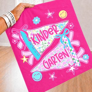 Peut inclure: Un carr&eacute; de tissu rose vif pr&eacute;sentant deux motifs en forme de fanion avec le texte "KINDER" et "GARTEN" en &eacute;criture rose. Les fanions sont d&eacute;cor&eacute;s de motifs floraux, de rubans bleus, d'&eacute;toiles jaunes et d'ic&ocirc;nes de visages souriants.