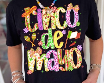 May include: Black t-shirt with a colorful "Cinco de Mayo" graphic. The design features a sombrero, maracas, and the Mexican flag. The text is in various patterns and colors, including leopard print and floral designs.