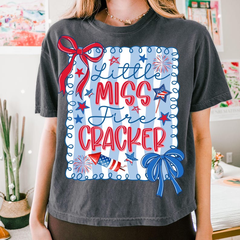 May include: Dark grey t-shirt with a patriotic design. The shirt features a rectangular graphic with the words "Little Miss Fire Cracker" in red and blue, along with stars, fireworks, and bows. The design is set against a blue and white striped background.