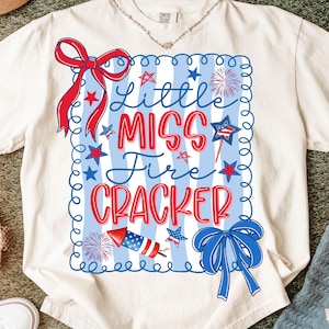 May include: Cream-coloured t-shirt with a patriotic design. The shirt features the text "Little Miss Fire Cracker" in red and blue, surrounded by stars, fireworks, and bows. The shirt is made of a soft material.