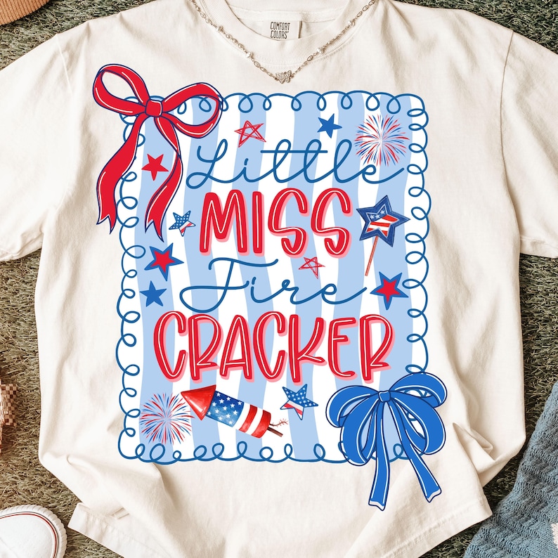 May include: Cream-coloured t-shirt with a patriotic design. The shirt features the text "Little Miss Fire Cracker" in red and blue, surrounded by stars, fireworks, and bows. The shirt is made of a soft material.