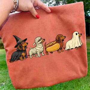 Peut inclure: Un sac fourre-tout couleur terre cuite avec une impression graphique de quatre teckels en costumes d'Halloween : un sorcier avec un chapeau et une cape noirs, un momifi&eacute;, un d&eacute;guis&eacute; en hot-dog et un portant un drap de fant&ocirc;me blanc.