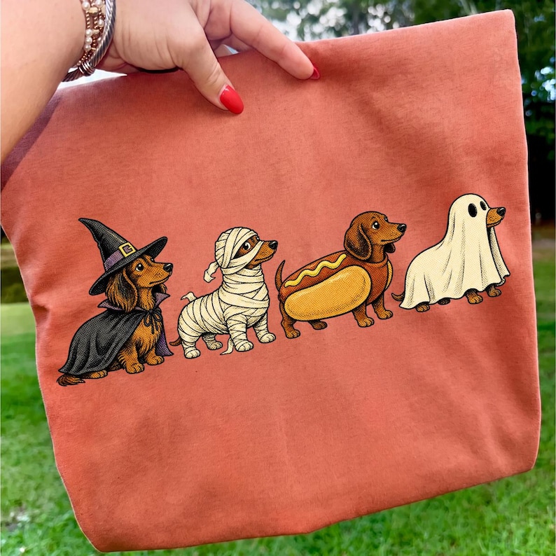 Peut inclure: Un sac fourre-tout couleur terre cuite avec une impression graphique de quatre teckels en costumes d'Halloween : un sorcier avec un chapeau et une cape noirs, un momifi&eacute;, un d&eacute;guis&eacute; en hot-dog et un portant un drap de fant&ocirc;me blanc.
