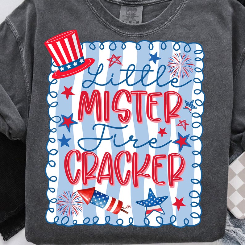 May include: Dark grey sweatshirt with a patriotic design. The design features a red, white, and blue top hat, stars, and the text "Little Mister Fire Cracker" in red and blue. A rocket and fireworks are also included.