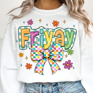 Peut inclure: Sweat-shirt blanc avec un motif &agrave; carreaux color&eacute;s qui dit "Friyay" avec un n&oelig;ud &agrave; carreaux arc-en-ciel. Le motif est entour&eacute; de petites fleurs et d'&eacute;toiles.