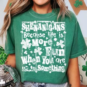 May include: Green t-shirt with the words "Shenanigans Because life is More Fun When You are up to Something" in white. The design includes shamrocks and stars. The shirt is a crew neck and has short sleeves.
