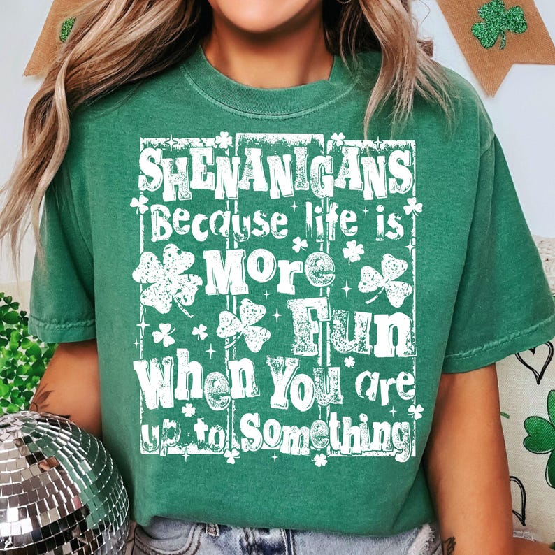 May include: Green t-shirt with the words "Shenanigans Because life is More Fun When You are up to Something" in white. The design includes shamrocks and stars. The shirt is a crew neck and has short sleeves.