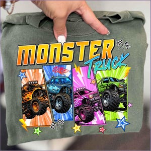 May include: Olive green t-shirt featuring the words "MONSTER TRUCK" in orange and blue. The shirt displays four colorful monster truck illustrations with flames, lightning bolts, and checkered flags. The design is set against a comic book style background.