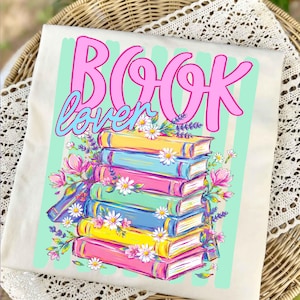 Puede incluir: Una camiseta color crema con un gr&aacute;fico de una pila de libros coloridos y el texto "BOOK lover" en rosa y azul. El dise&ntilde;o incluye ilustraciones de margaritas blancas, ramas de lavanda moradas y flores rosas sobre rayas de color verde menta.