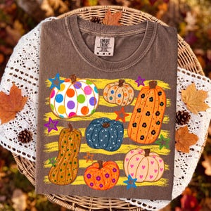May include: A brown crew neck t-shirt with a Comfort Colors label at the collar. The front features a graphic of six pumpkins in various shapes and colors, including polka dots, floral patterns, and hearts, accented by several colorful stars.