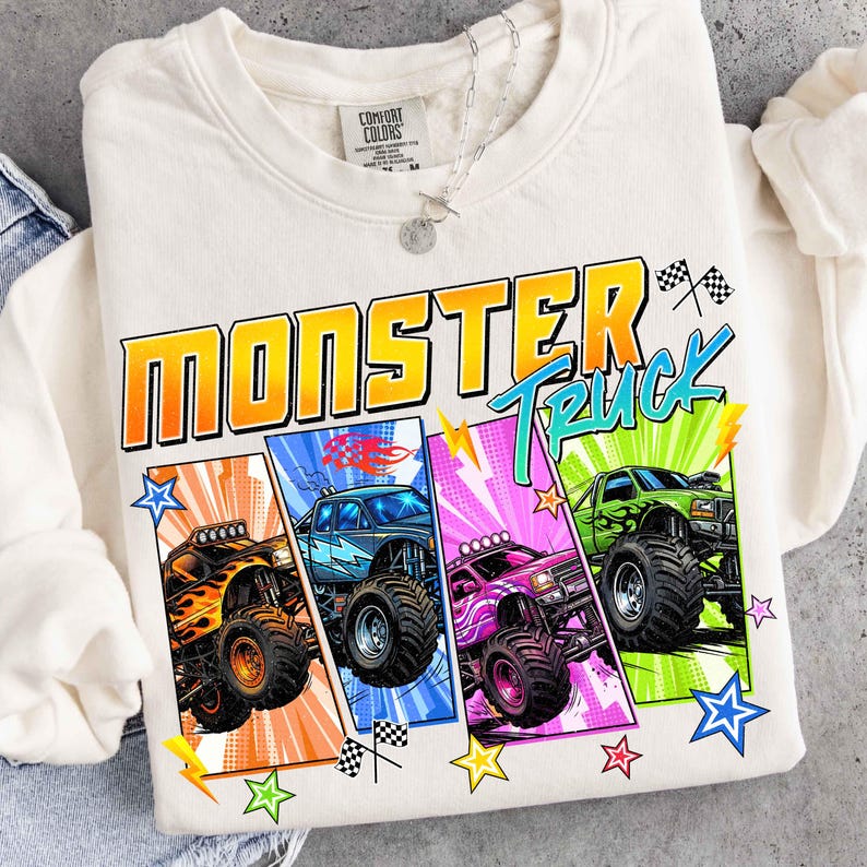 May include: Cream-colored sweatshirt featuring a colorful graphic of four monster trucks. The design includes the words "MONSTER TRUCK" in orange and teal, along with checkered flags and stars. The trucks are in orange, blue, pink, and green.
