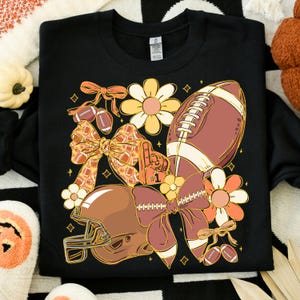 May include: Black sweatshirt with a football-themed design. The graphic includes a football, helmet, bows, flowers, and a foam finger. The design features shades of brown, orange, and yellow.
