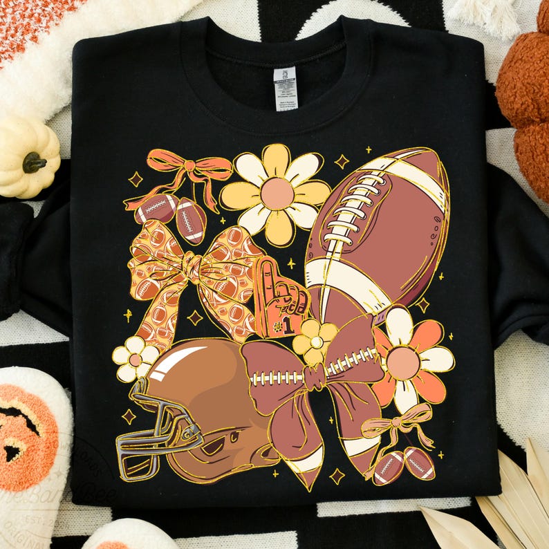 May include: Black sweatshirt with a football-themed design. The graphic includes a football, helmet, bows, flowers, and a foam finger. The design features shades of brown, orange, and yellow.