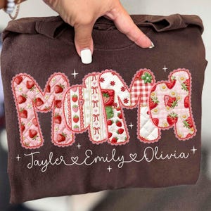 May include: A brown sweatshirt with the word "MAMA" in a patchwork design featuring strawberries, gingham, and floral patterns. The names "Tayler", "Emily", and "Olivia" are written below the word "MAMA".