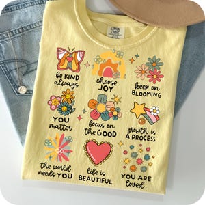 May include: Yellow t-shirt with a colorful graphic design featuring flowers, butterflies, and inspirational messages like "be kind always", "choose joy", "keep on blooming", "you matter", "focus on the good", "growth is a process", "the world needs you", "life is beautiful", and "you are loved".