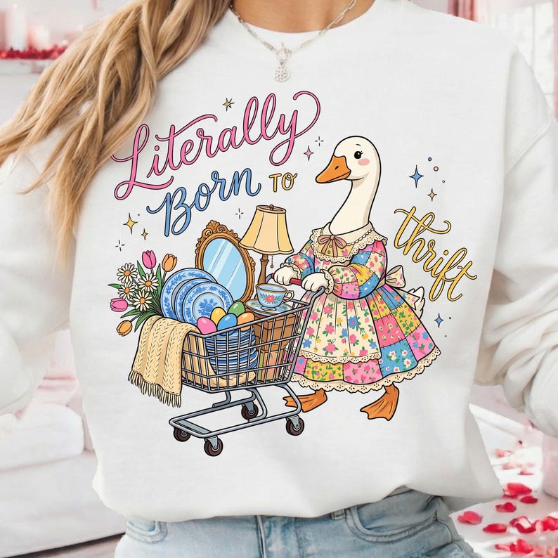Może przedstawiać: Biała bluza z zabawną ilustracją gęsi pchającej w&oacute;zek sklepowy wypełniony przedmiotami vintage. Tekst "Literally Born to Thrift" jest napisany kolorową czcionką. Gęś ma na sobie patchworkową sukienkę.
