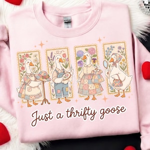 Pode incluir: Camisola rosa com uma ilustração fantasiosa de quatro gansos vestidos com roupas de patchwork, cada um emoldurado com bordas florais. O texto "Just a thrifty goose" está escrito em uma fonte de script na parte inferior. A camisola tem decote redondo.