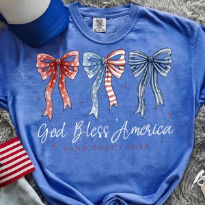 May include: A blue t-shirt with three bow designs in red, white, and blue, with stars and stripes. The text "God Bless America" and "Land That I Love" are printed below the bows. A blue baseball cap is visible in the upper left corner.